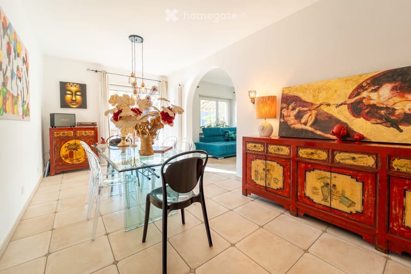 Dining area with a glass table, chairs, a sofa in the background, decorative cabinets, a lamp, and artwork on the walls.