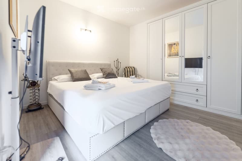 Modern bedroom with a large white bed, pillows, a wardrobe, a chair, a screen, a table, and a rug.