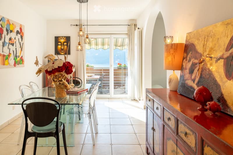 Dining room with a glass table, chairs, a decorative vase, a wooden cabinet, and artwork on the walls. Large windows offer a view of the sea.