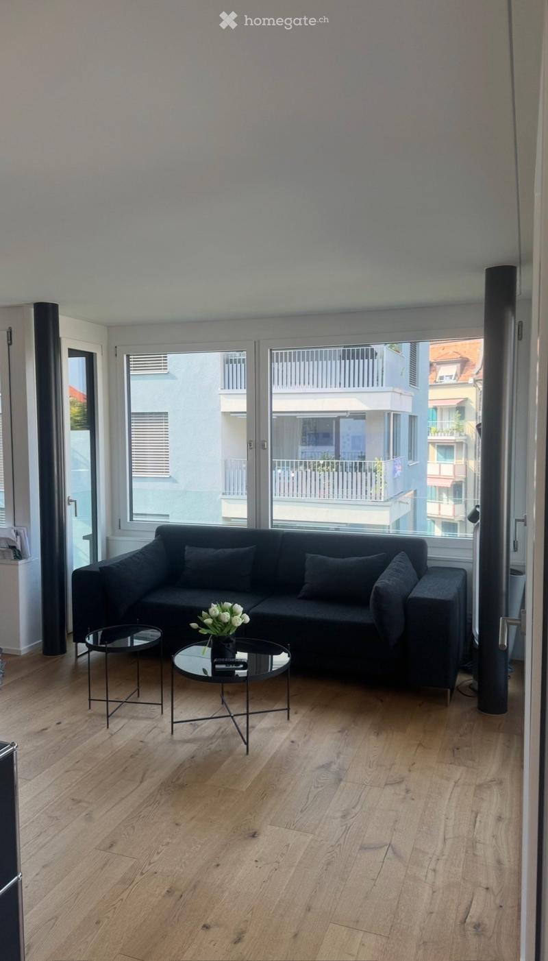 Modern living room with light wooden flooring, large windows, black sectional sofa, two round glass coffee tables, and a flower vase on one of the tables.