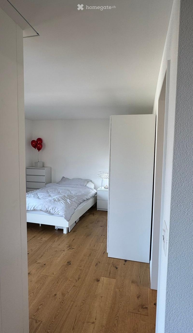 Minimalist bedroom with a white bed, nightstand, lamp, and balloons on a wooden floor.