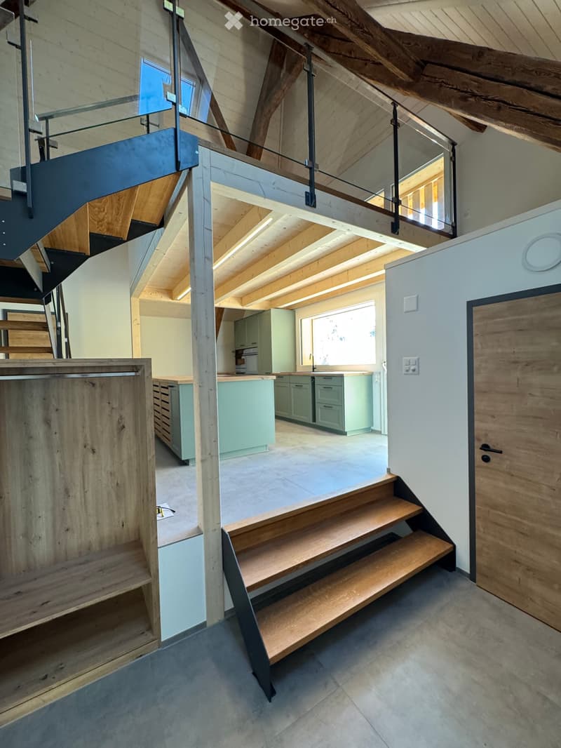 Modern open-concept hallway with wooden staircase, glass railing, and visible kitchen area with light green cabinets and island.