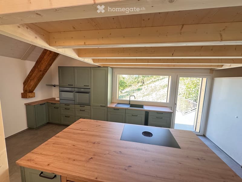 Modern kitchen with light green cabinets, wooden island, built-in oven, sink, and induction cooktop. Large window and open door leading to outdoor area.