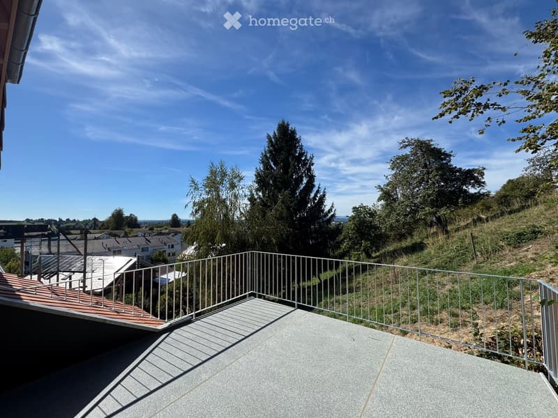 A modern balcony with a metal railing, concrete floor, and a view of a green, hilly landscape with trees and houses in the distance.