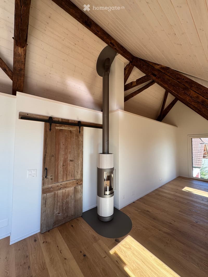 An empty attic space with exposed wooden beams, a modern wood-burning stove, and a large window letting in natural light.