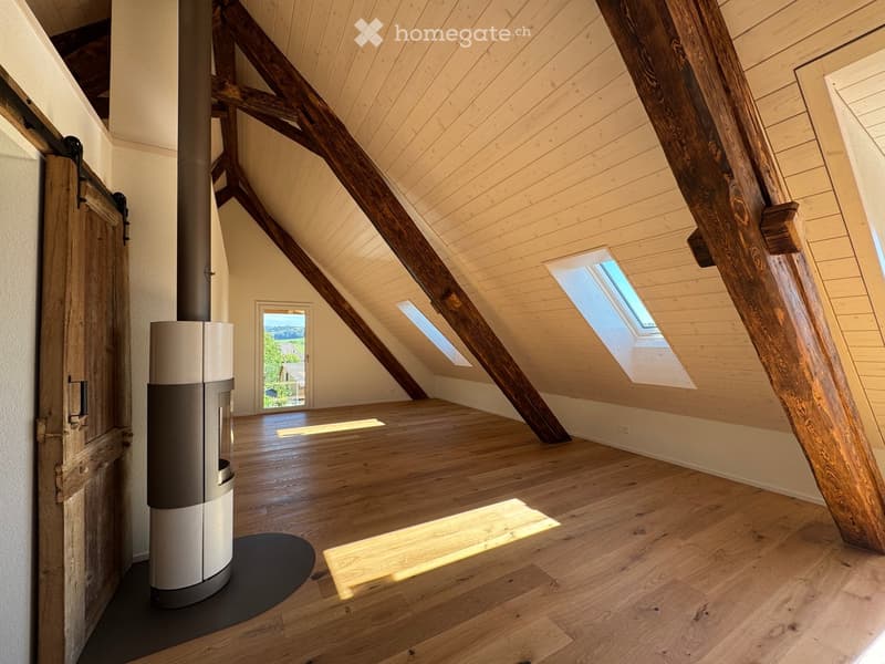 Bright, airy attic space with wooden beams, skylights, and a modern wood-burning stove. The floor is made of light wood planks, and the walls are painted in a soft, neutral tone.