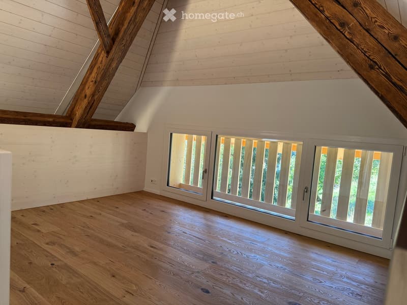 Empty attic room with wooden floors, white paneled walls, exposed wooden beams, and large windows with white frames showing greenery outside.
