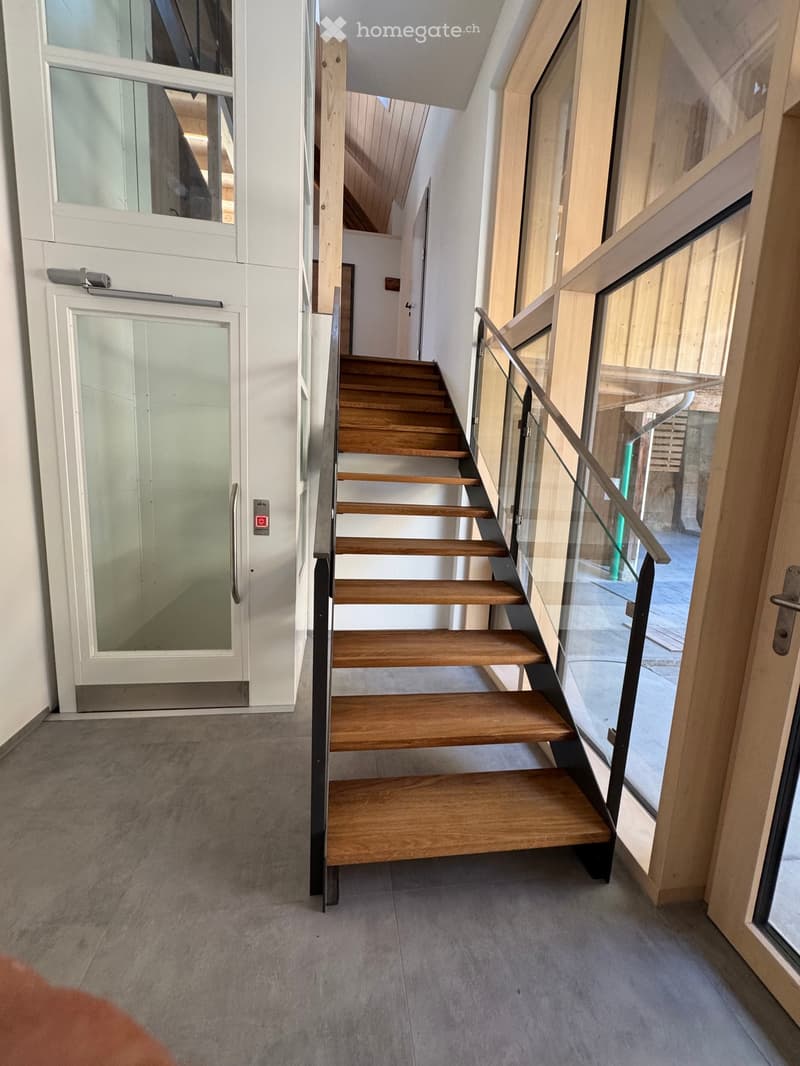 Modern hallway with wooden staircase, glass railing, and elevator access.