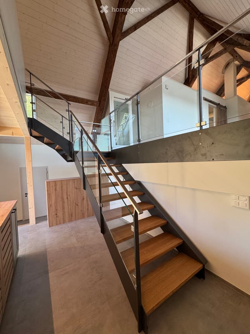 Modern staircase with wooden steps and metal railing, glass balustrade, exposed wooden beams, and a raised ground floor visible in the background.