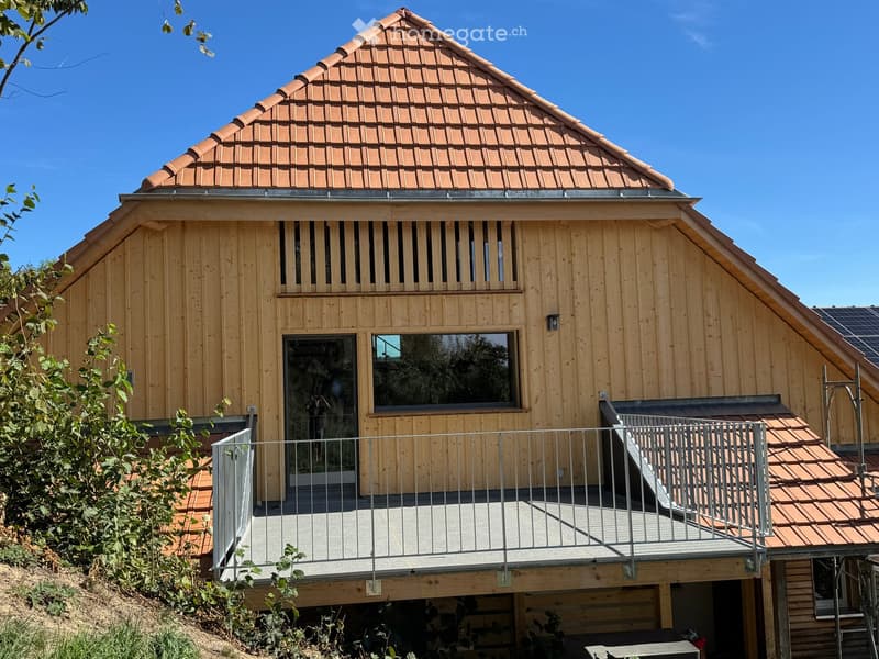 Modern wooden house with a sloped red-tiled roof, large windows, and a raised balcony with metal railings.