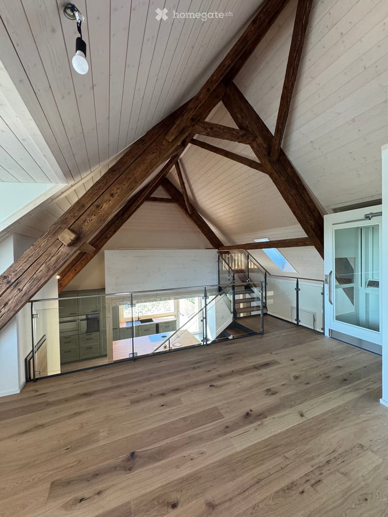 An attic space with exposed wooden beams and a glass staircase leading to a lower level. The floor is made of polished hardwood, and the ceiling features a skylight and pendant light fixture.