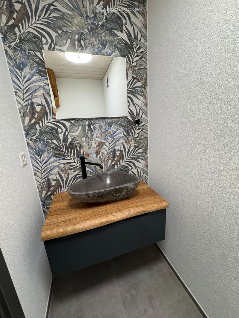 Modern bathroom with a wooden countertop, stone sink, black faucet, and a built-in cabinet below. The walls are decorated with a leafy, tropical print.
