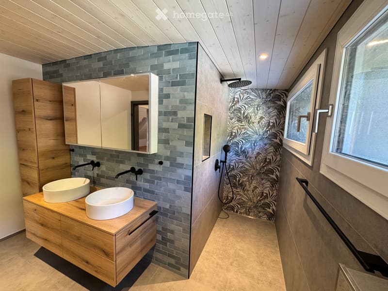 Modern bathroom with wooden vanity, two sinks, large mirror, and a shower area with a decorative wall.