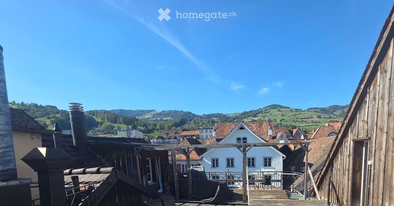 Balcony with wooden decking, overlooking a village with traditional houses and green hills in the background. Includes a table and chairs.