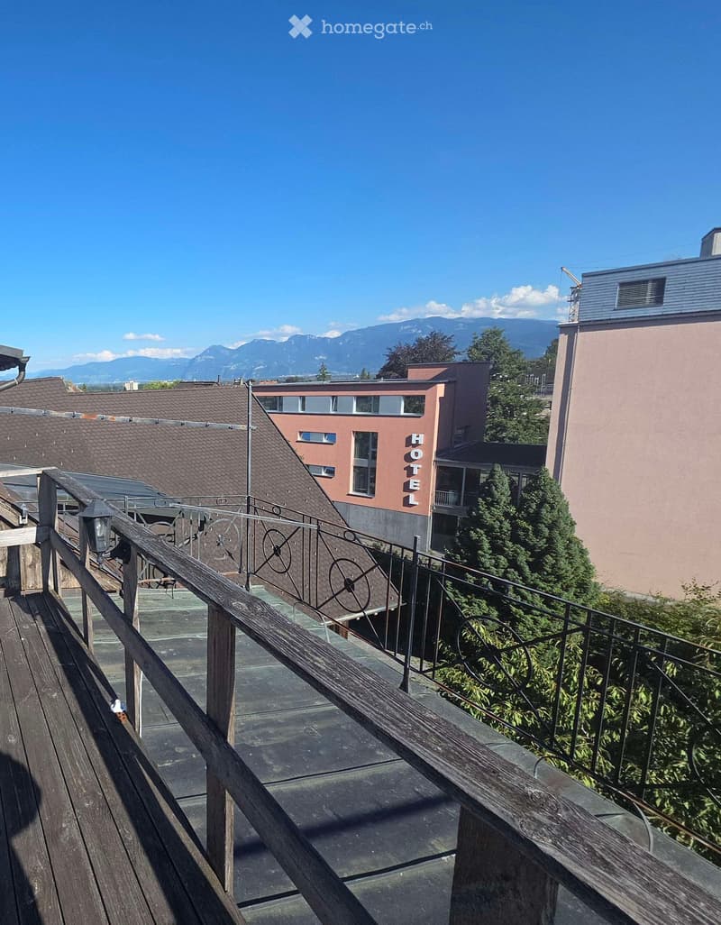 Balcony with wooden decking, metal railing, and steps leading down, overlooking a hotel building and surrounding landscape with mountains in the background.