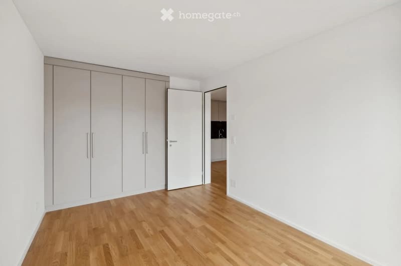 Empty room with light wooden flooring, white walls, and built-in white closet units. An open doorway leads to another room with a visible kitchen area.