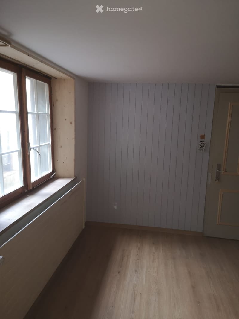 Empty room with wooden flooring, light grey paneled walls, and a single window with wooden frame and sill.