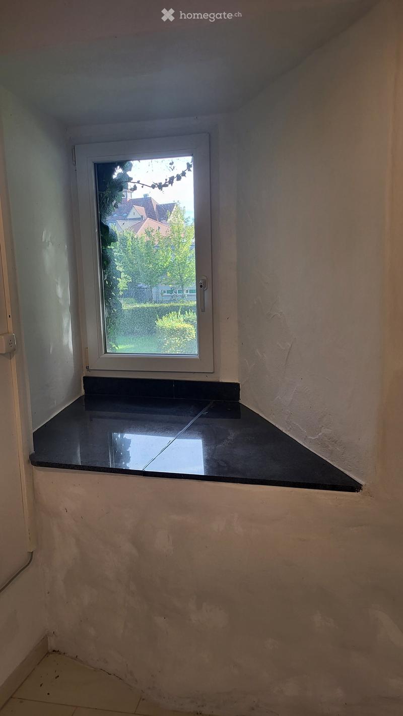 Empty room with a window and a small black countertop below it. The walls are plain and the floor is tiled.