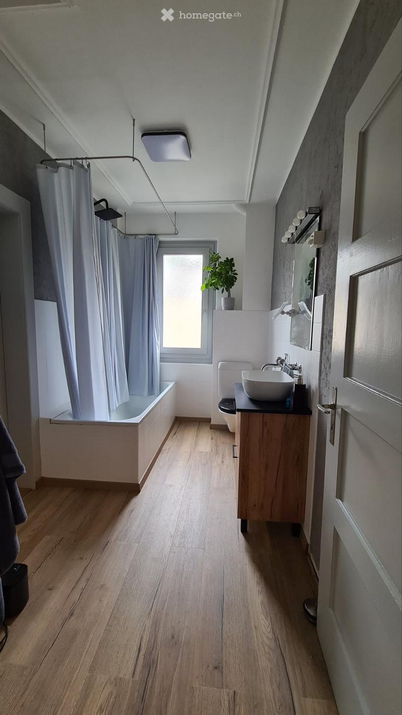 Modern bathroom with wooden flooring, a bathtub, a sink with a faucet, a mirror, and a small plant on the windowsill.
