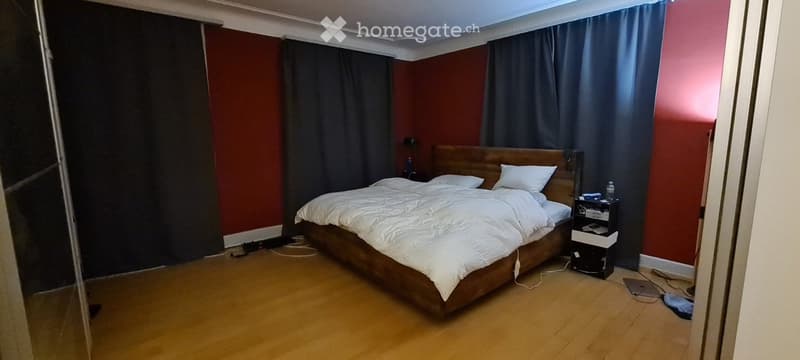 Bedroom with wooden floor, red walls, large window with dark curtains, bed with white bedding, nightstand with items, and visible cables on the floor.