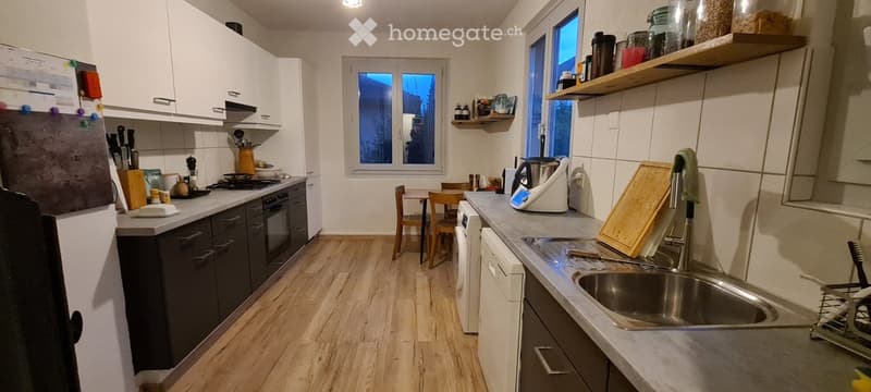 Modern kitchen with white cabinets, dark countertops, and wooden flooring. Features include a stainless steel sink, refrigerator, oven, and various kitchen appliances and utensils.