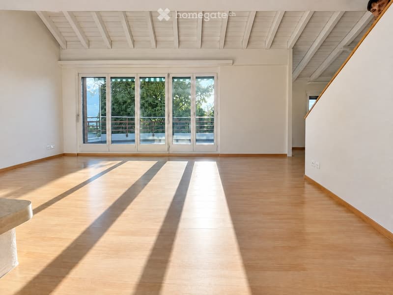 Bright, empty room with wooden flooring, white walls, and large glass doors leading to a balcony. The ceiling has exposed wooden beams.