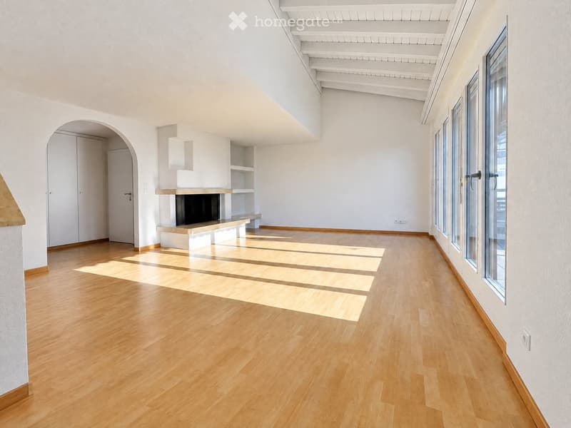 Empty room with light wooden flooring, white walls, and large windows allowing natural light. Features include a fireplace with built-in shelving and an arched doorway leading to another area.