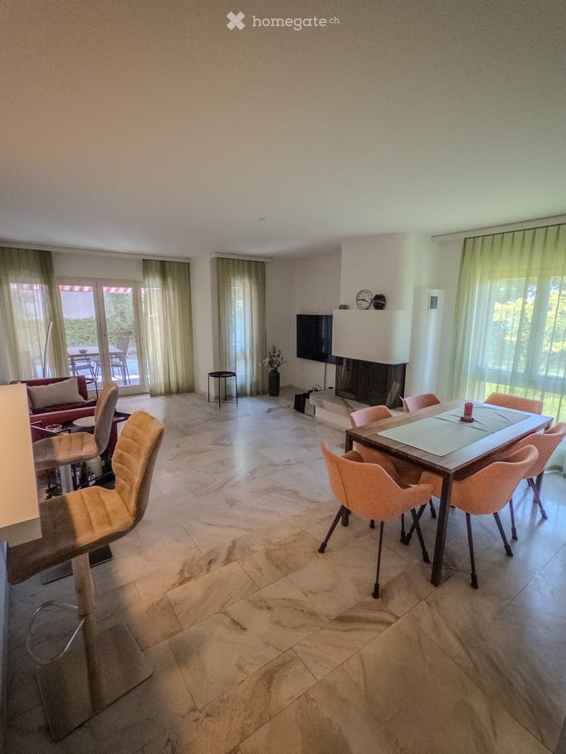 Bright living room with marble flooring, a fireplace, a sofa, a dining table with chairs, and a television mounted on the wall. Large windows with sheer curtains let in natural light.