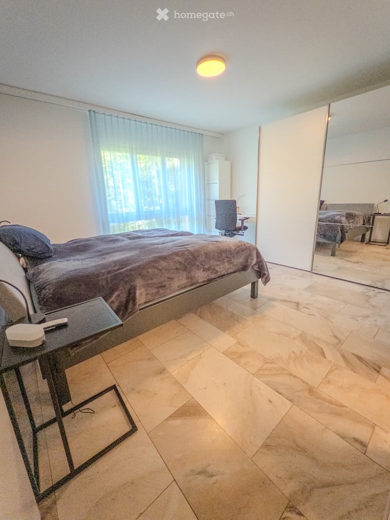 Modern bedroom with a large bed, nightstand, chair, and mirror. The room features large windows with sheer curtains and a tiled floor.