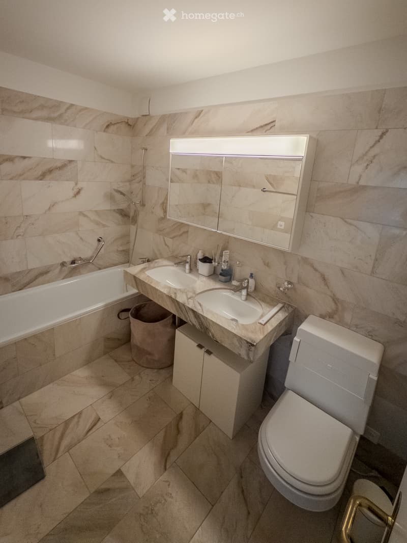 Modern bathroom with marble tiles, double sink, bathtub, toilet, and built-in mirror cabinet.