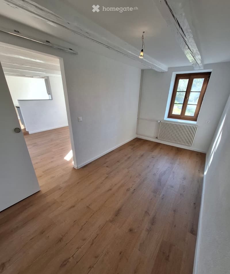 Empty room with wooden flooring, white walls, and a single window with wooden frame. Exposed wooden beams on the ceiling and a radiator under the window.