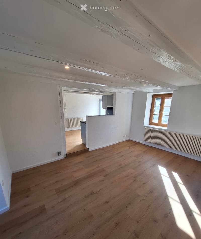 Empty room with wooden flooring, white walls, and a window with wooden frame. The room has exposed wooden beams on the ceiling and a radiator under the window.