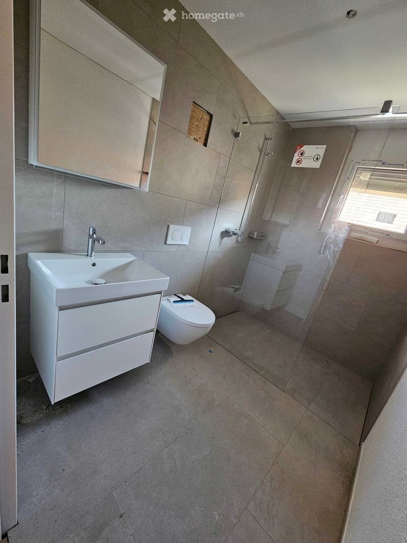 Modern bathroom with a white sink, faucet, cabinet, and toilet. The walls are tiled, and the floor is made of light-colored tiles.
