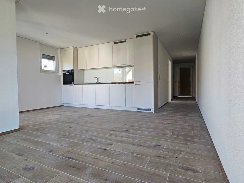 Empty hallway with wooden flooring, white walls, and a small kitchen area visible at one end. The kitchen features white cabinets, a countertop, and a built-in oven.