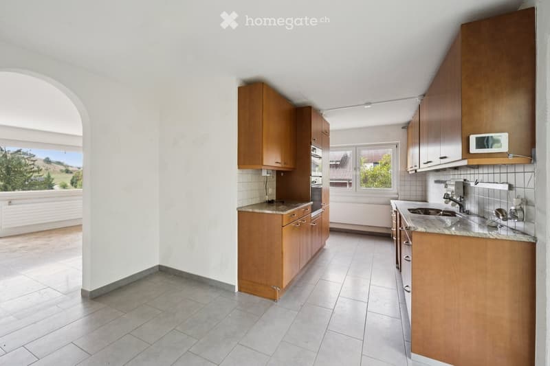 Bright kitchen with wooden cabinets, granite countertops, and tiled floors. Features an oven, sink, and faucet, with large windows offering a scenic view.