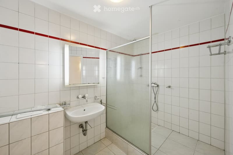 Modern bathroom with white tiles, a red accent border, a glass-enclosed shower, a white sink with a mirror above it, and a towel rack.