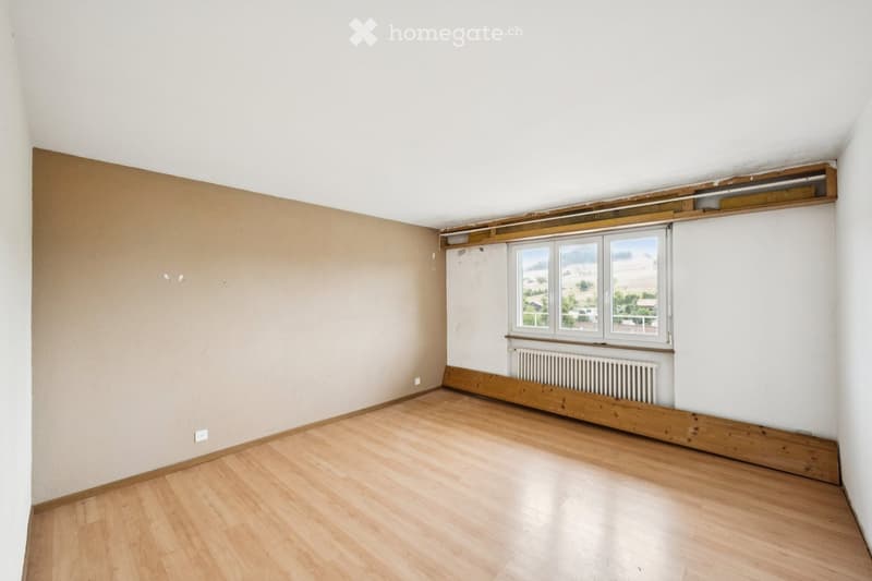 Empty room with wooden flooring, white walls, and a large window offering a scenic view. The room features a radiator below the window and a small wooden shelf above it.