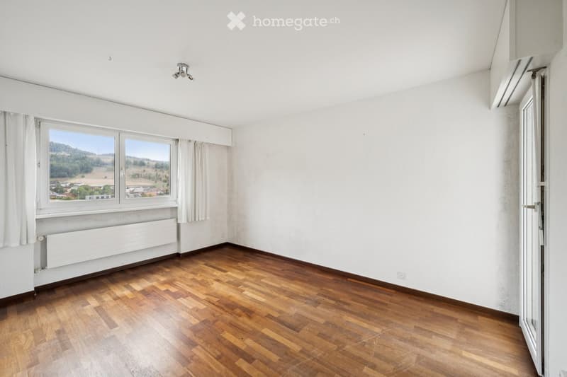Empty room with wooden flooring, white walls, large windows with white curtains, and a radiator under the windows.