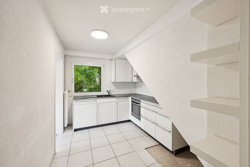 Modern, minimalist kitchen with white cabinets, a sink, and an oven. The space features a large window with a view of greenery outside, and the ceiling has a recessed light fixture.