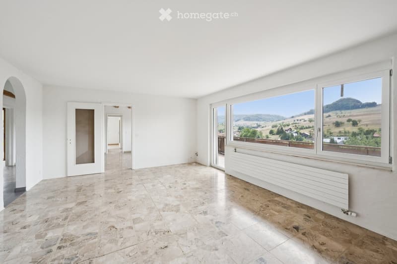 Empty room with white walls, tiled floor, large windows offering a scenic view, and a radiator below the windows.
