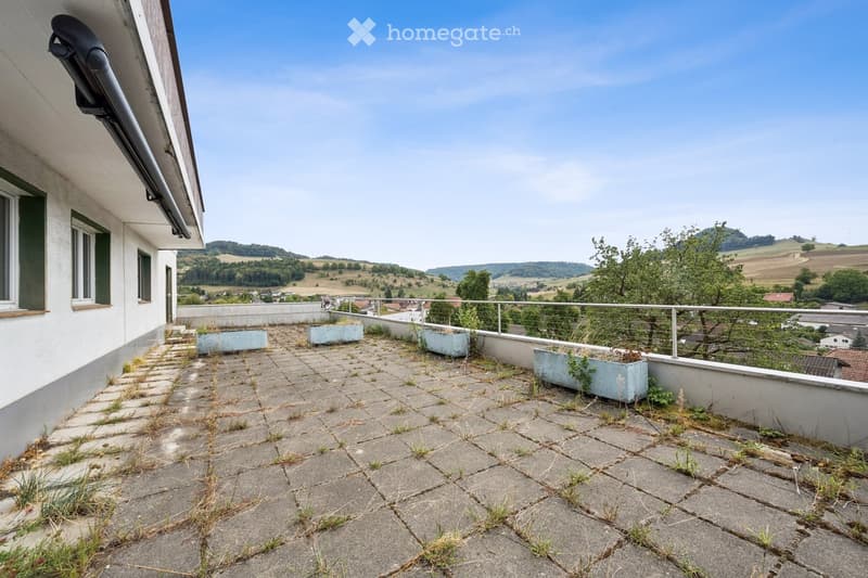 Roof terrace with concrete tiles, metal railing, and a few rectangular planters, overlooking a rural landscape with hills and scattered buildings.