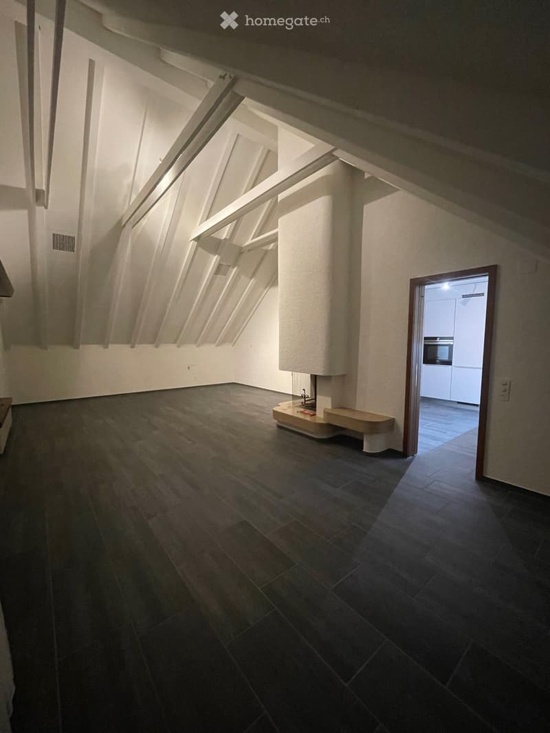 Empty attic space with wooden flooring, exposed wooden beams, and a small built-in bench near the wall. A doorway leads to another room with white cabinets and a built-in oven.