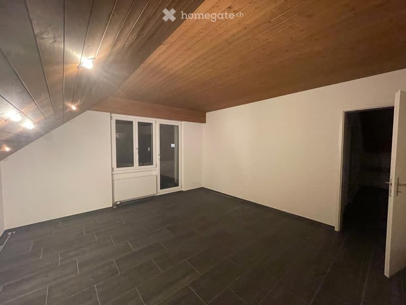 Empty room with dark tiled floor, white walls, wooden slanted ceiling, and a large window with glass doors leading outside.