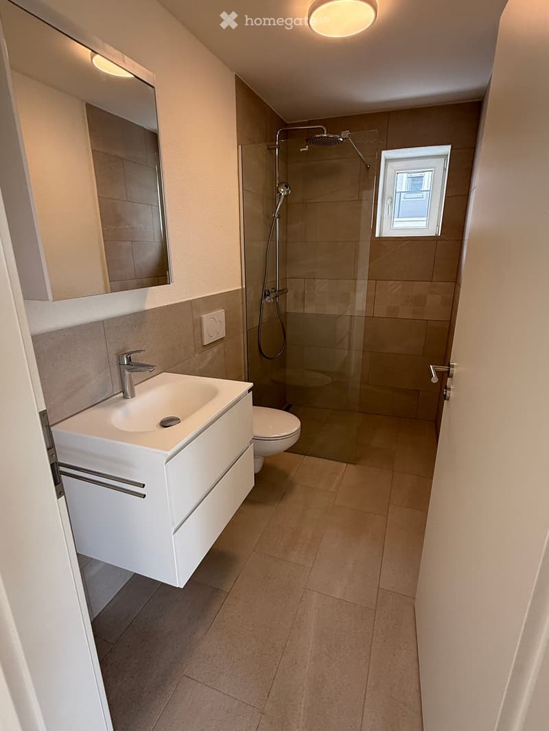 Modern bathroom with a white sink, mirror, toilet, and glass-enclosed shower. Features beige tiles and a small window.