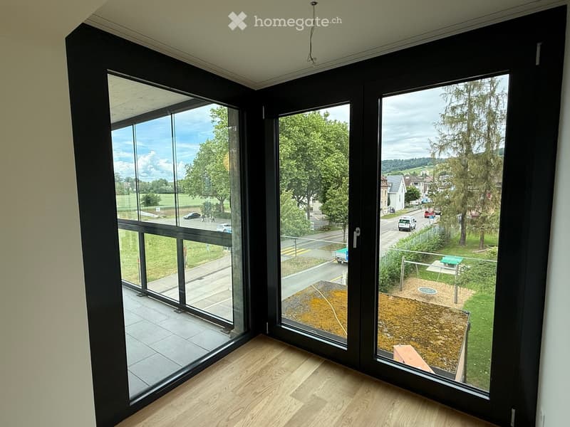 Large sliding glass doors open to a balcony with wooden flooring, overlooking a park and street with trees and buildings.
