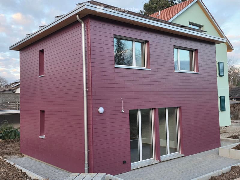 House. Two-story house with maroon siding, white-framed windows, and a modern design. Features include a flat roof section, a small dormer window, and a covered entrance area.