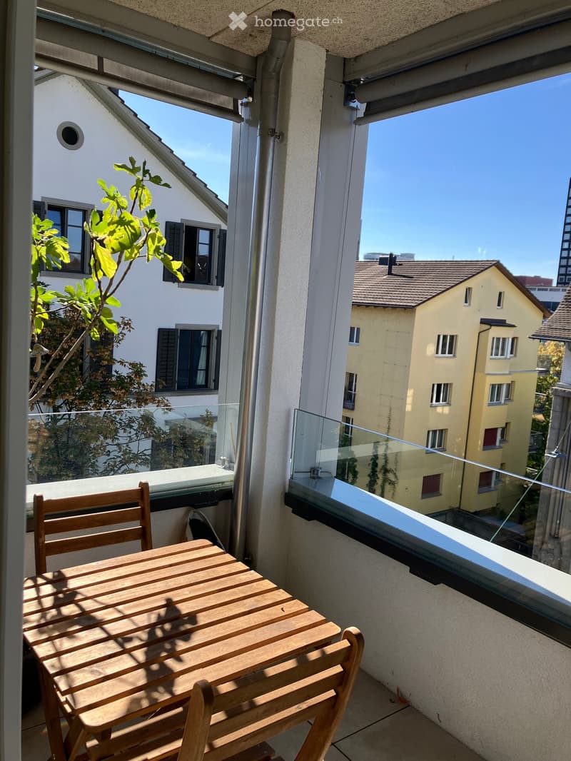 Balcony with a wooden table and chairs, glass railing, and a view of neighboring buildings.