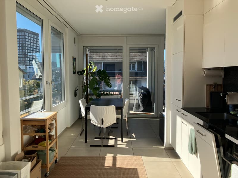 Bright, modern kitchen and dining area with white cabinets, black countertops, a sink, oven, dishwasher, a small cart, a dining table with chairs, and a large plant near the sliding glass doors.