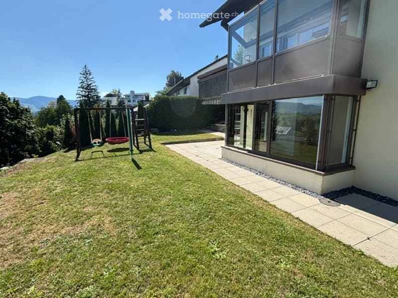 Well-maintained garden with a swing set, green grass, and a modern house with large windows and a view of the mountains.