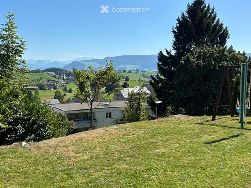 Lush green garden with a swing set, overlooking a scenic valley and distant mountains.